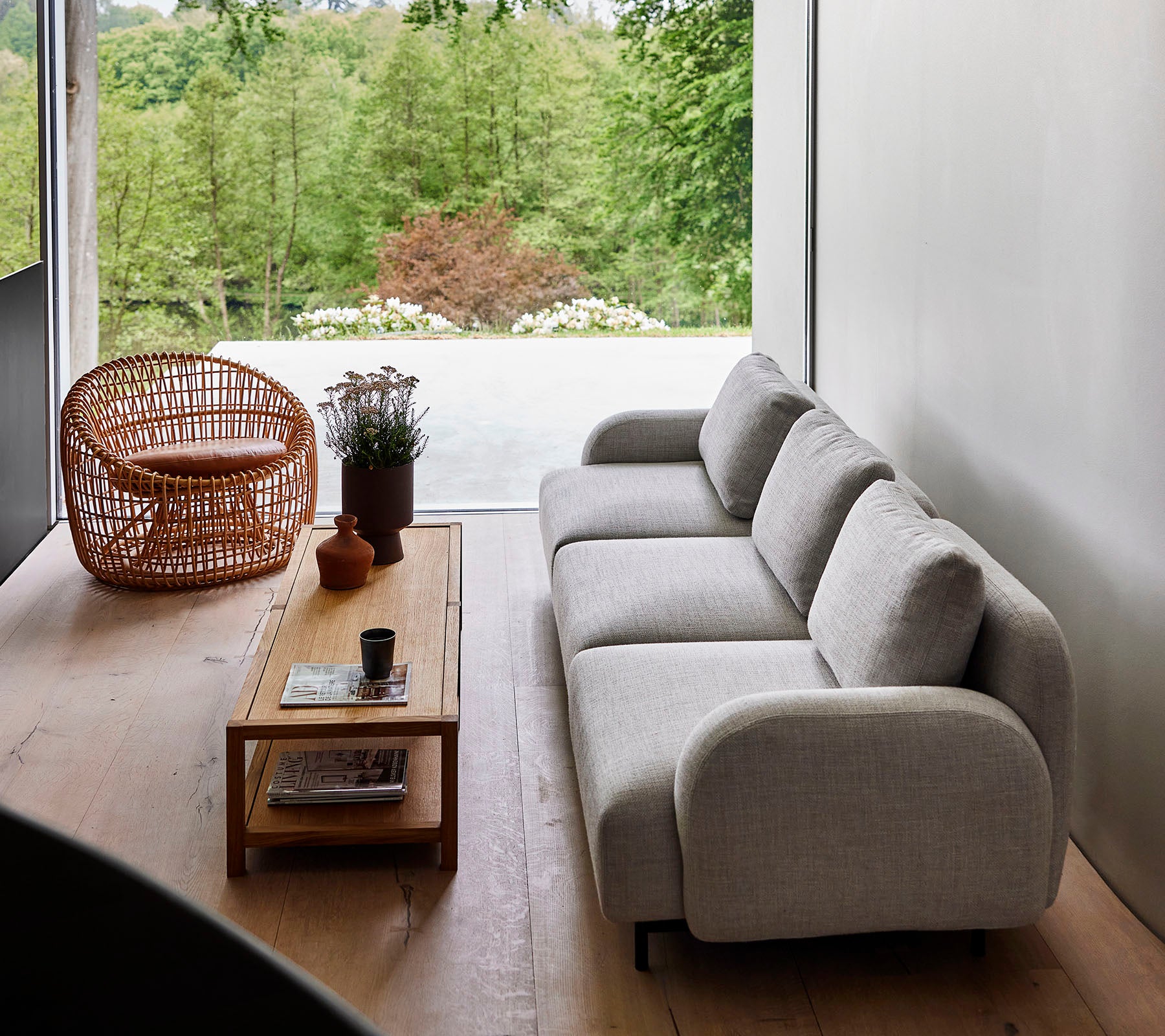 Modern sofa in light fabric paired with a wooden coffee table and wicker chair, surrounded by greenery. Natural sunlight enhances the serene ambiance.