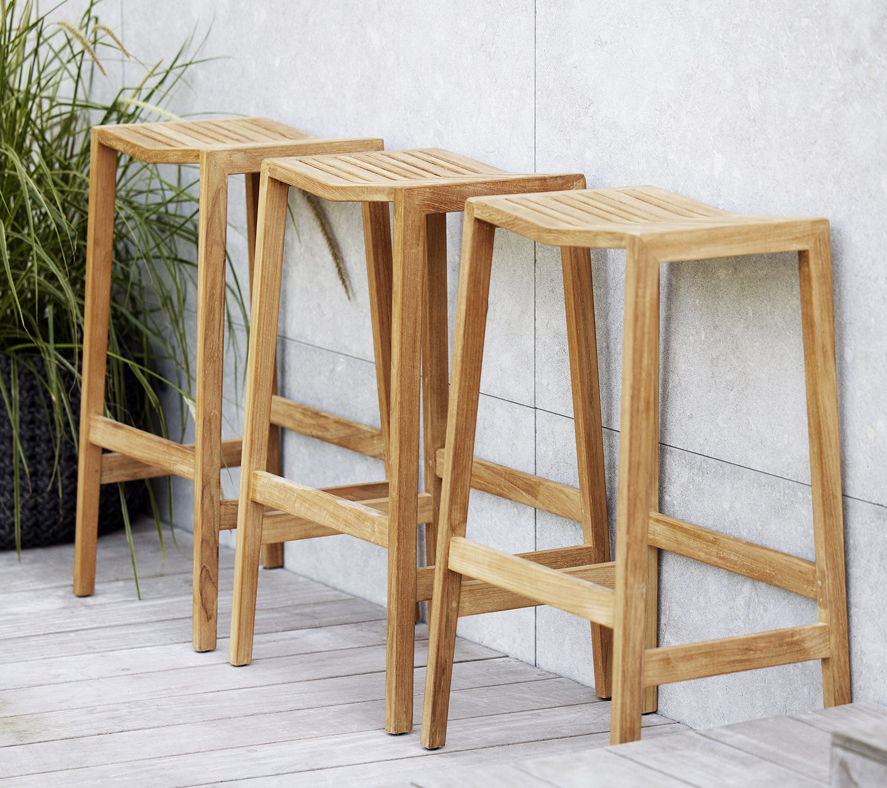 Three wooden flip bar stools with slatted seats, featuring a minimalist design and clean lines, positioned next to a decorative plant.