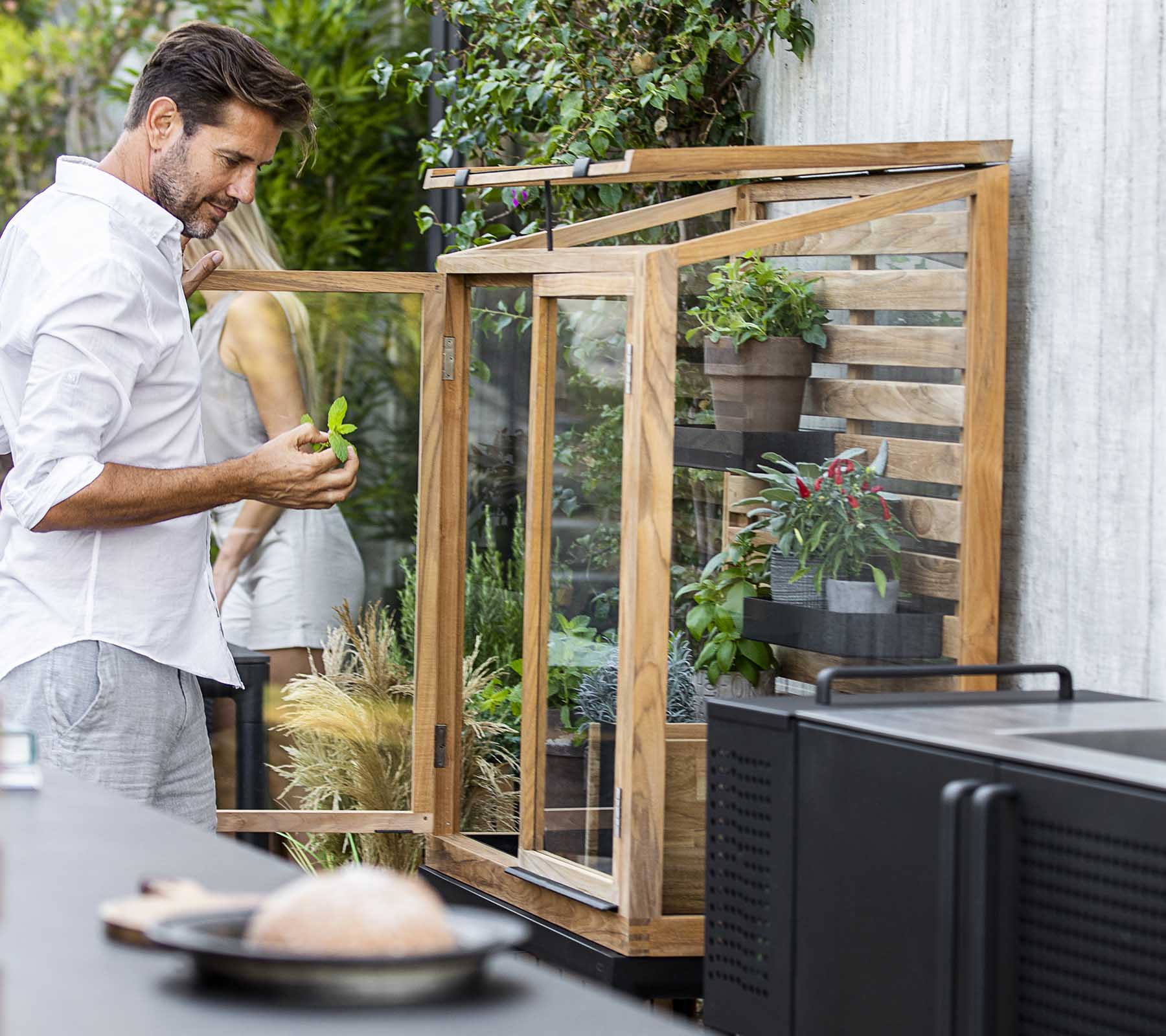 Man tending to plants in a glass display unit, surrounded by greenery, with a modern kitchen setup in the foreground.