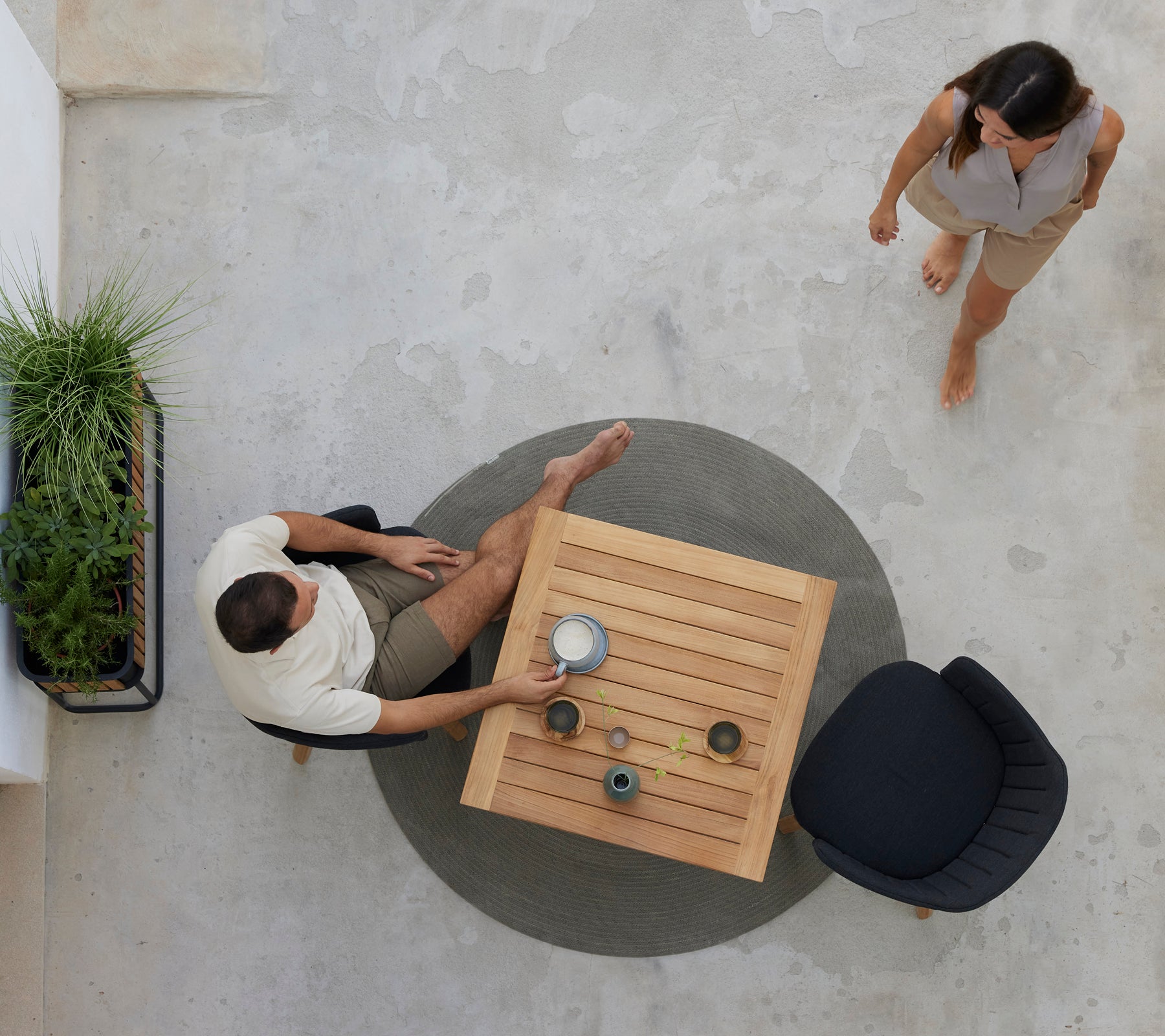 Two people enjoy coffee at a teak table surrounded by greenery, with a black chair nearby. A relaxed cafe atmosphere is portrayed.