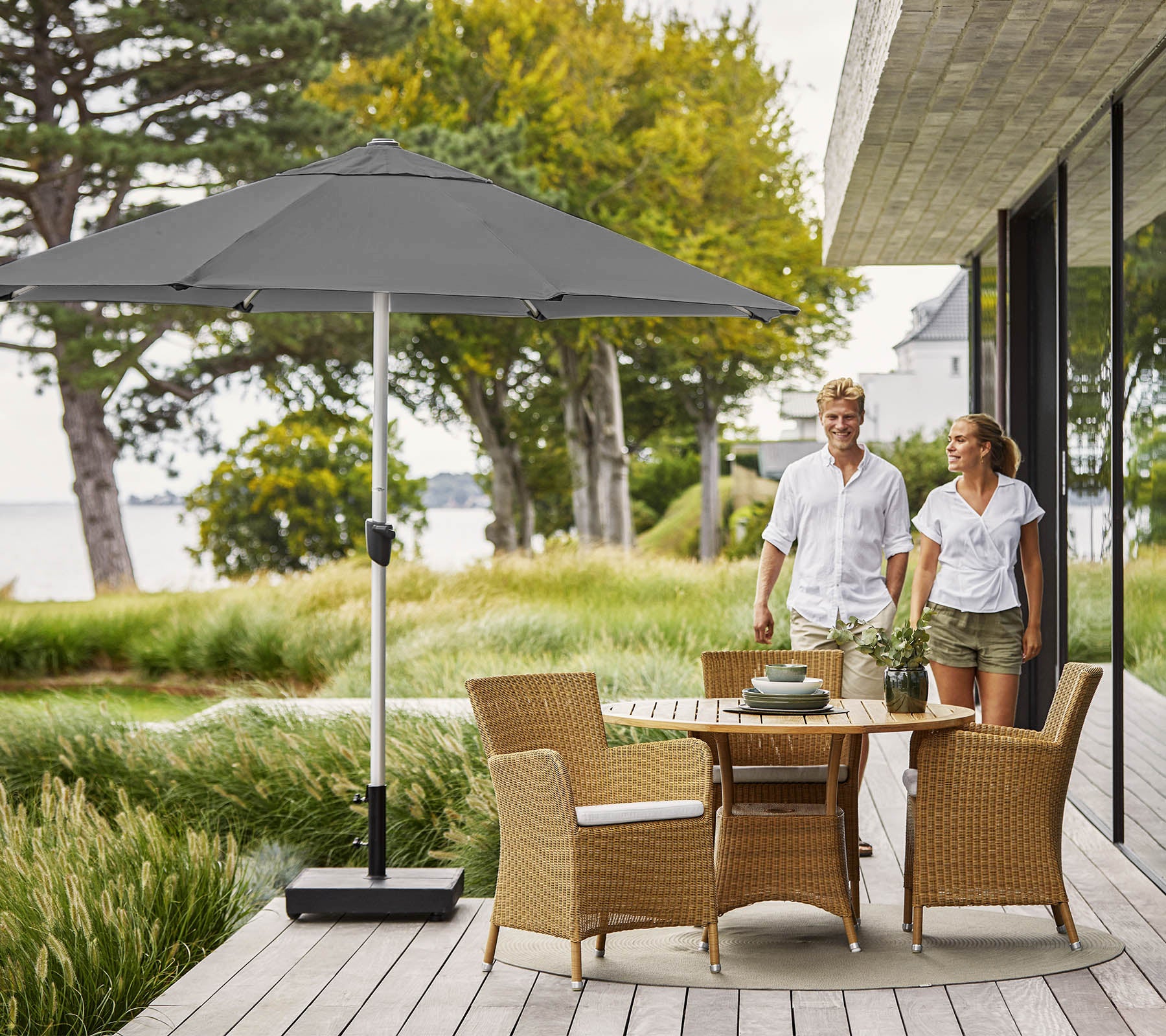 Couple walking on a wooden deck with a round table and wicker chairs, shaded by a large gray umbrella, overlooking lush greenery.
