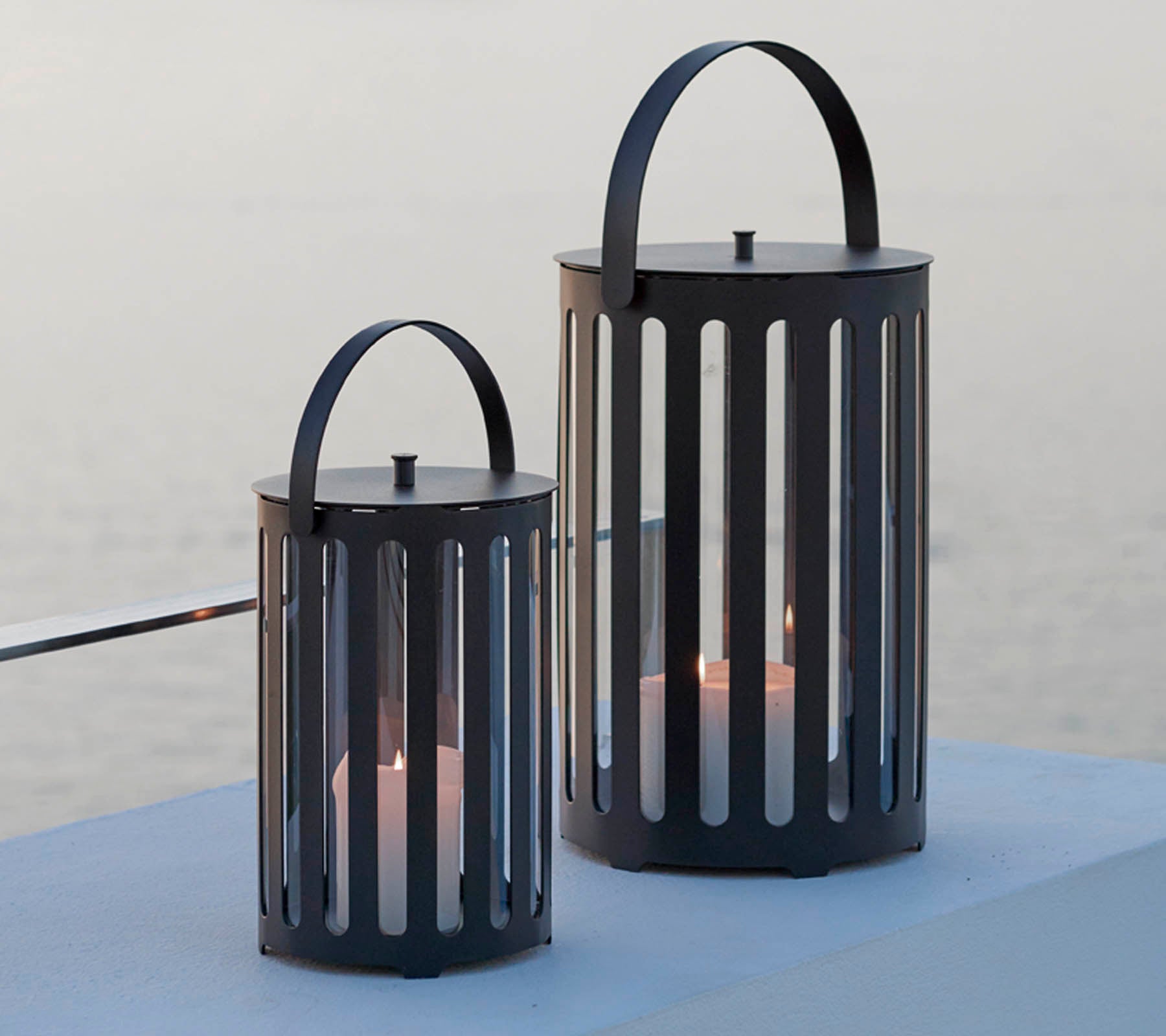 Two stylish metal lanterns with vertical slits, featuring handles and candles inside, set against a serene background.