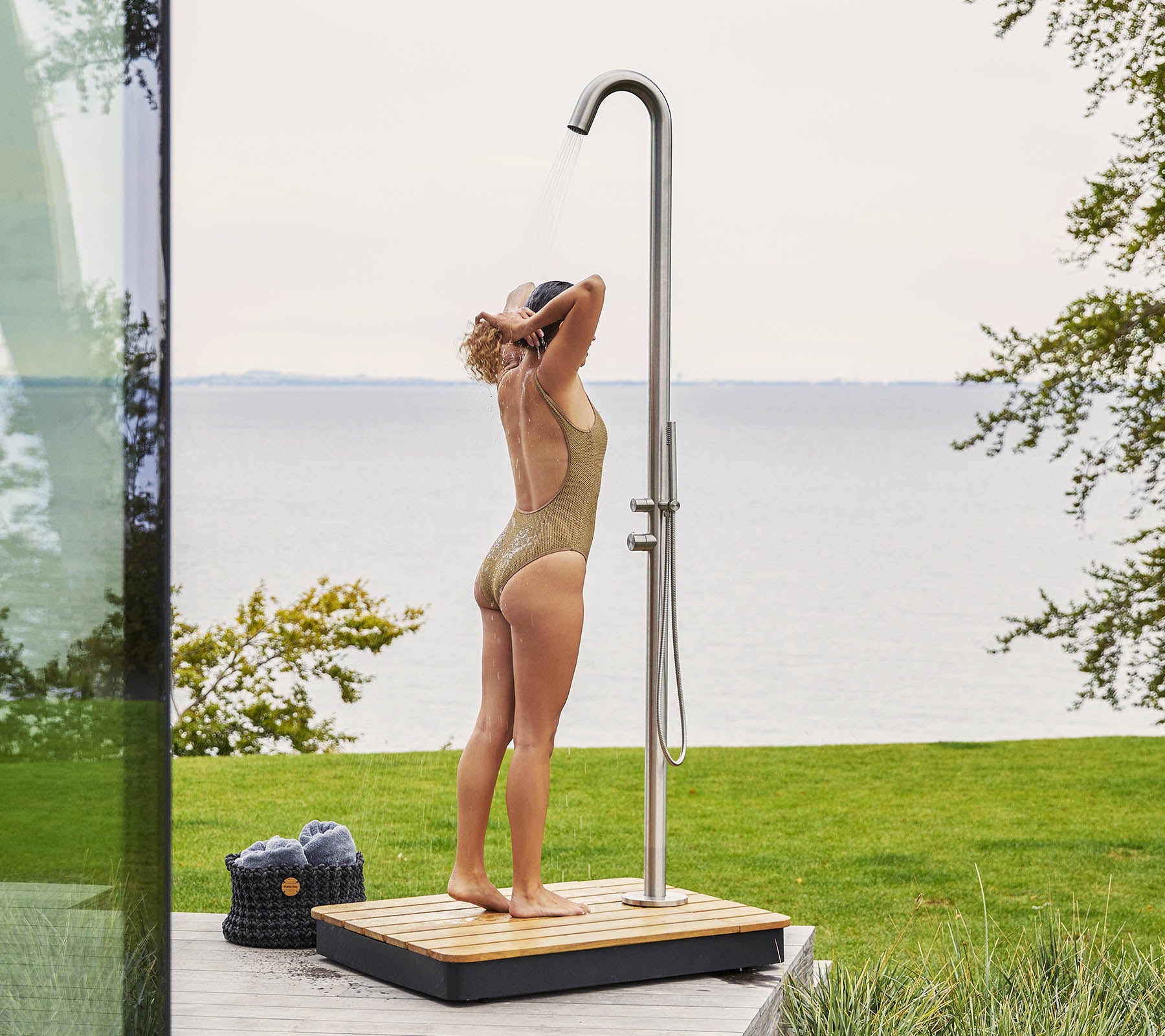 A person enjoying a refreshing outdoor shower, standing on a wooden base with water cascading from a tall, sleek showerhead.