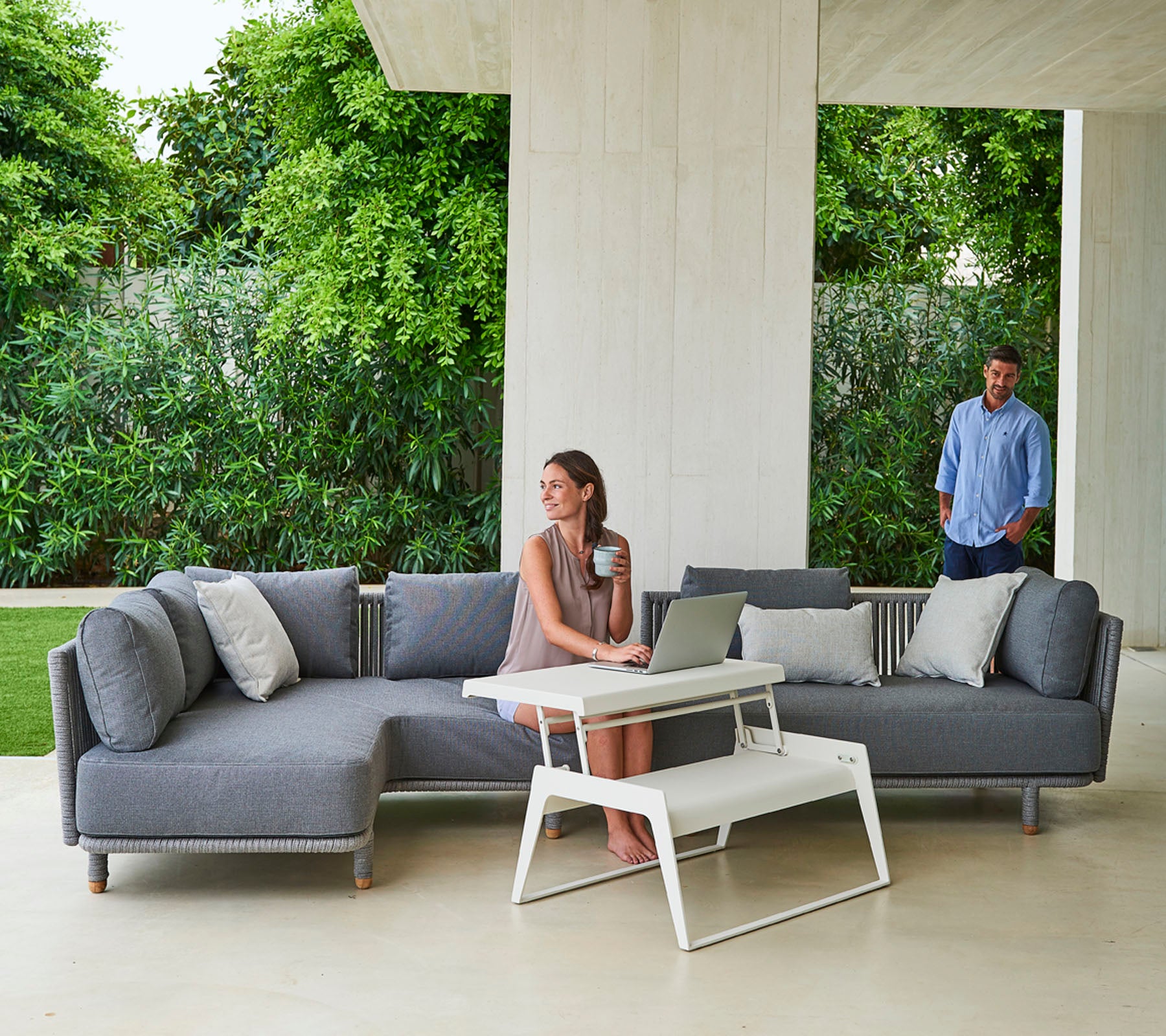 A stylish gray sectional sofa with cushions, accompanied by a modern coffee table, and a woman working on a laptop, while a man stands nearby.