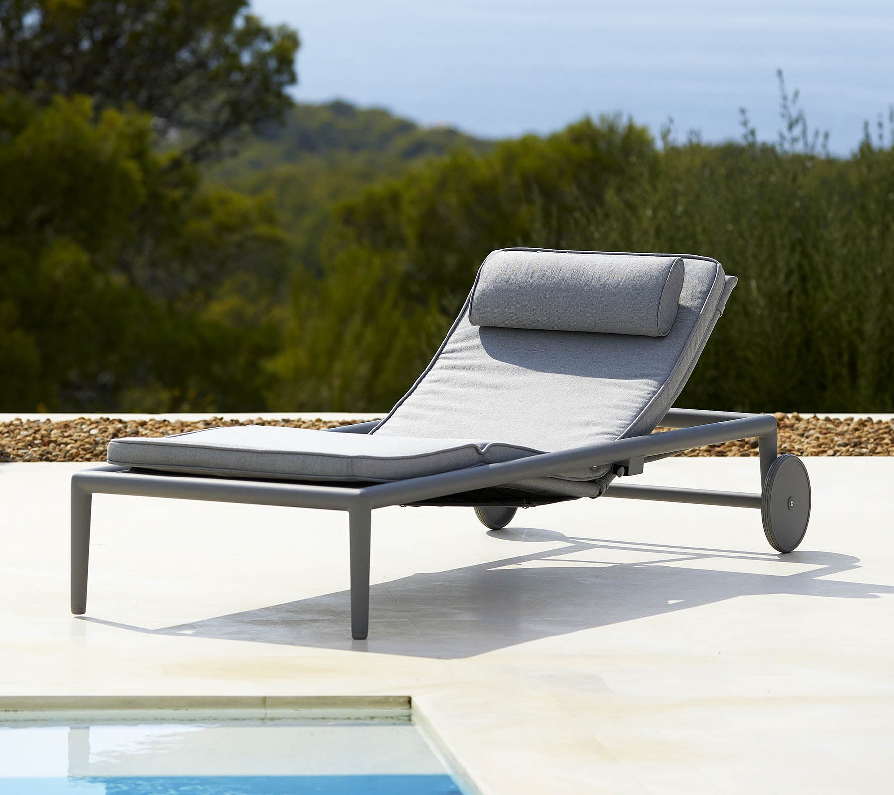 Comfortable lounge chair with a gray cushion and backrest, featuring wheels for easy mobility, beside a pool and lush greenery in the background.