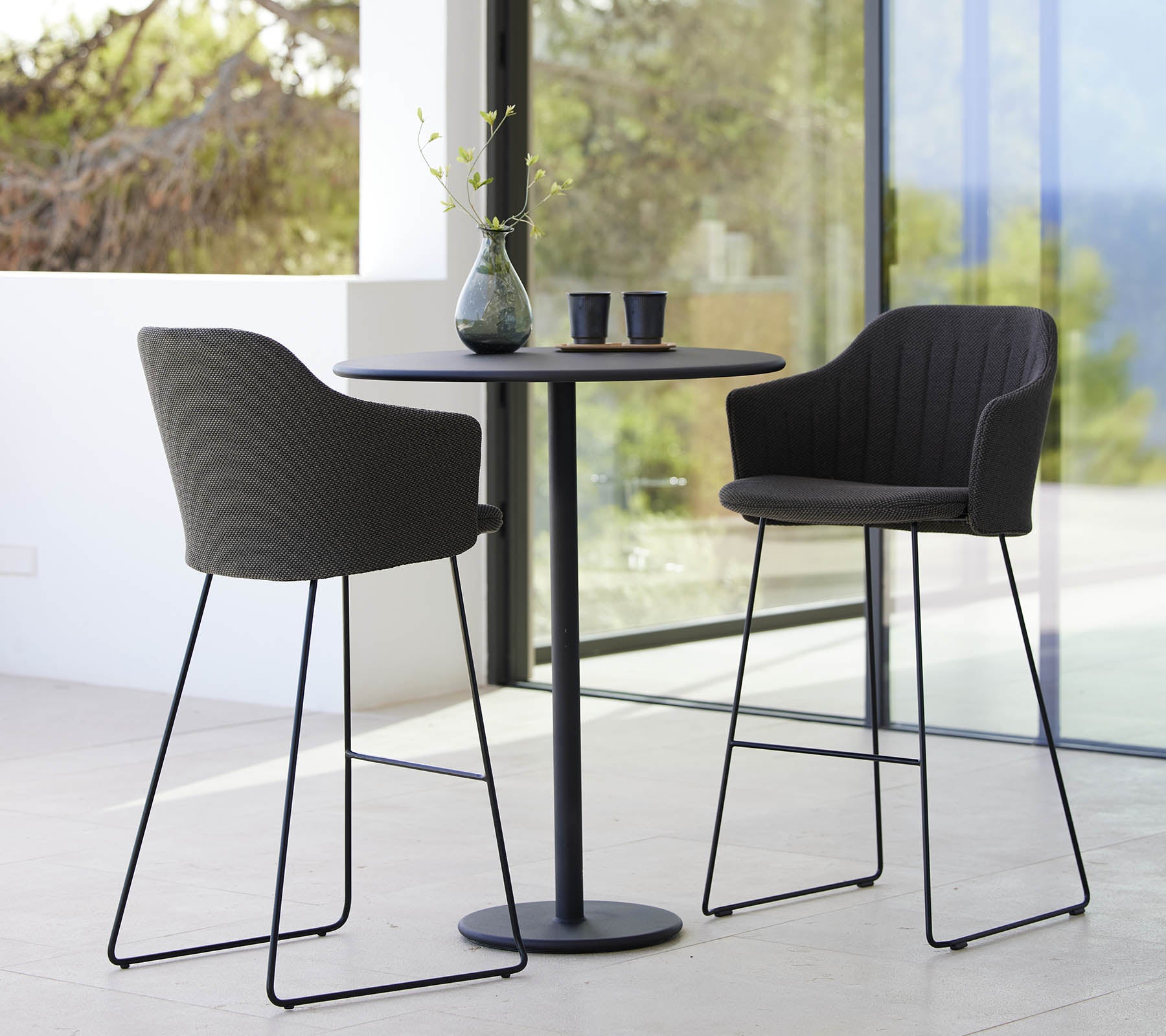 Two stylish chairs paired with a sleek bar table, complemented by a vase and cups, creating a modern and inviting seating arrangement.