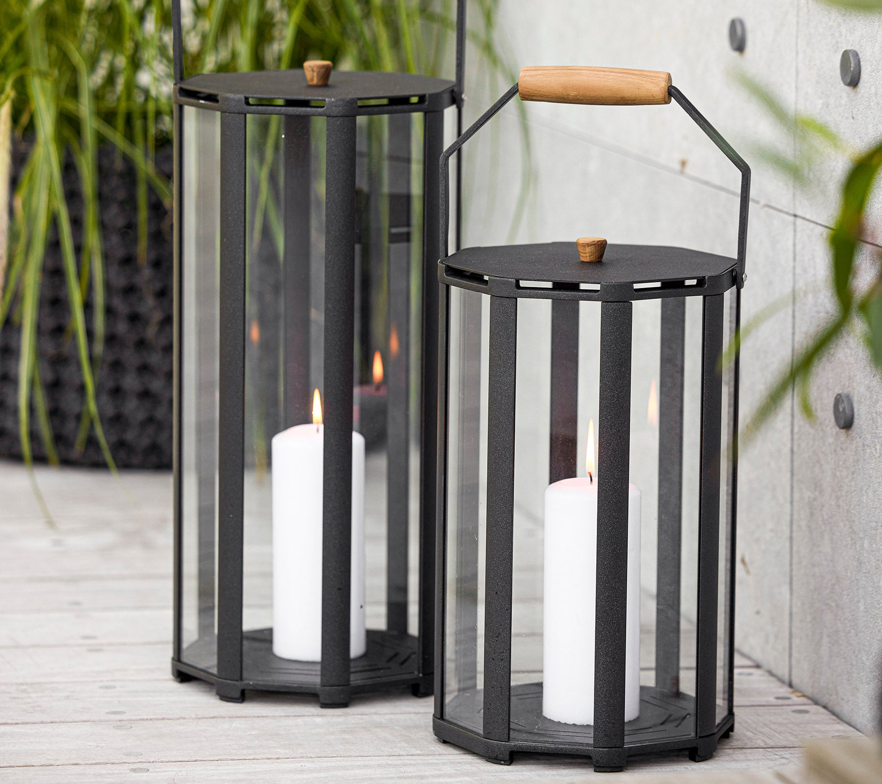 Two stylish lanterns with a modern design, featuring glass panels and a wooden handle, holding white candles inside.