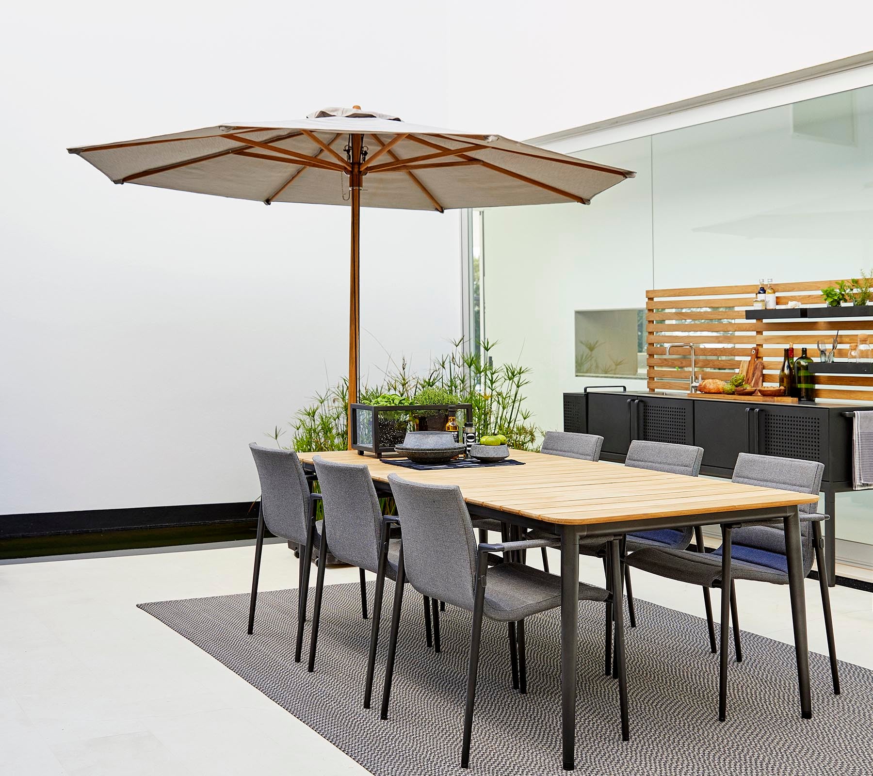 A classic dining set featuring a wooden table with an umbrella and gray chairs, surrounded by decorative plants on a stylish rug.
