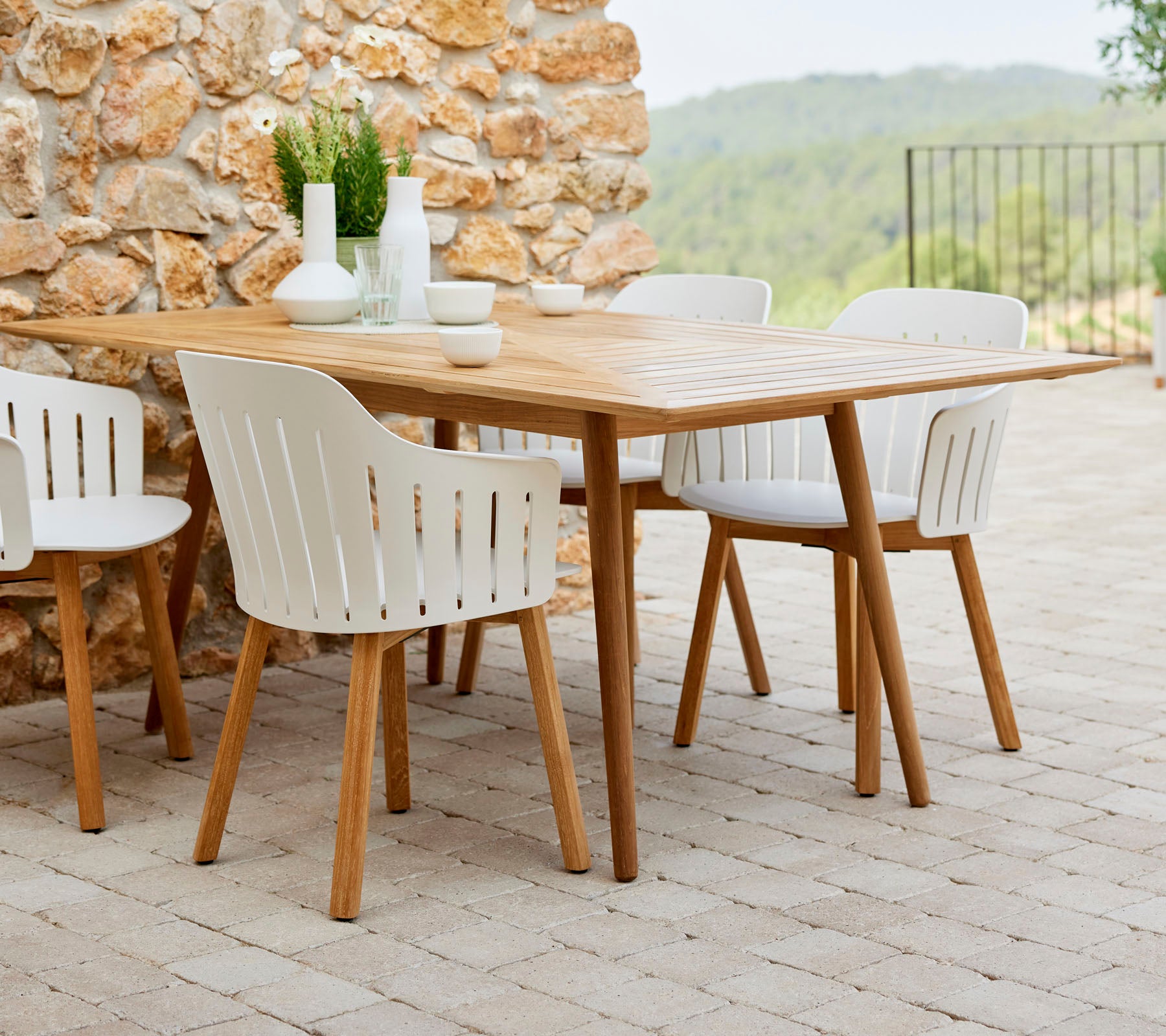 A wooden dining table surrounded by white chairs, complemented by decorative plants and simple tableware, set against a natural stone backdrop.