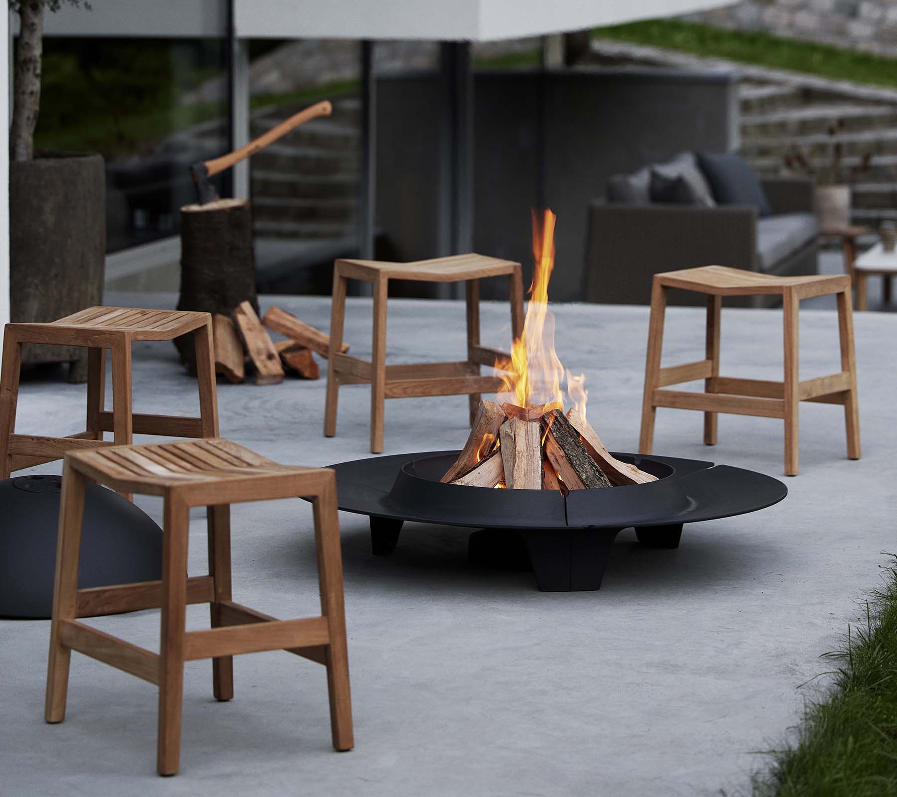 Wooden flip stools surround a circular ember fire pit, with a blazing fire at the center and logs stacked nearby.
