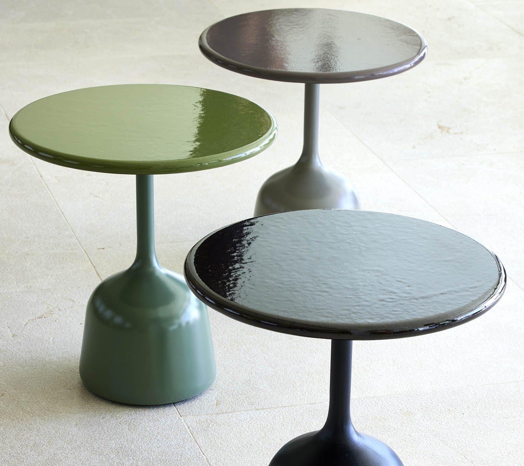 Three glazed coffee tables in green, black, and dark grey, each with a round top and a slender, curved base.