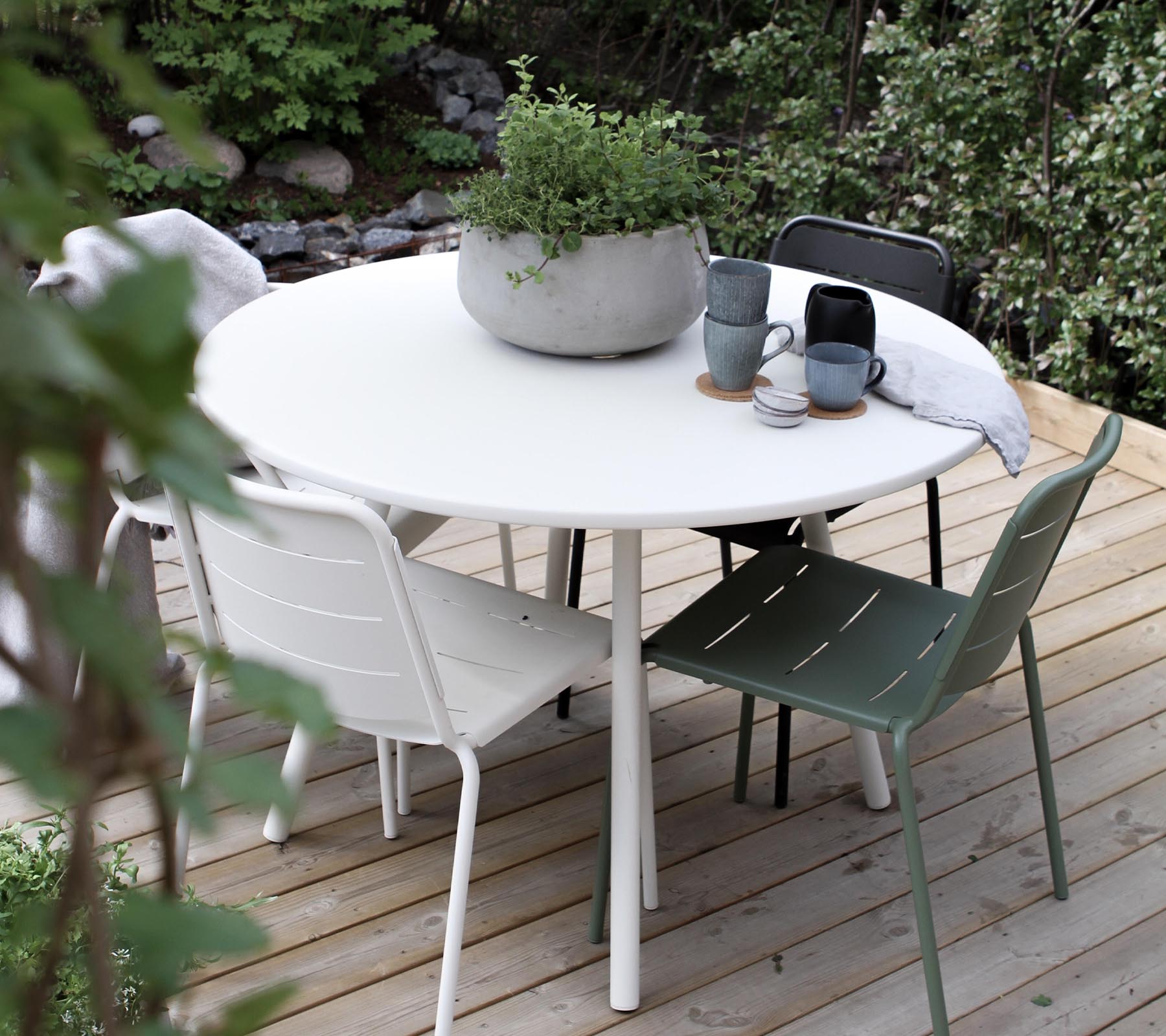 A round table surrounded by four chairs, featuring a potted plant and coffee mugs on the tabletop. Lush greenery in the background.