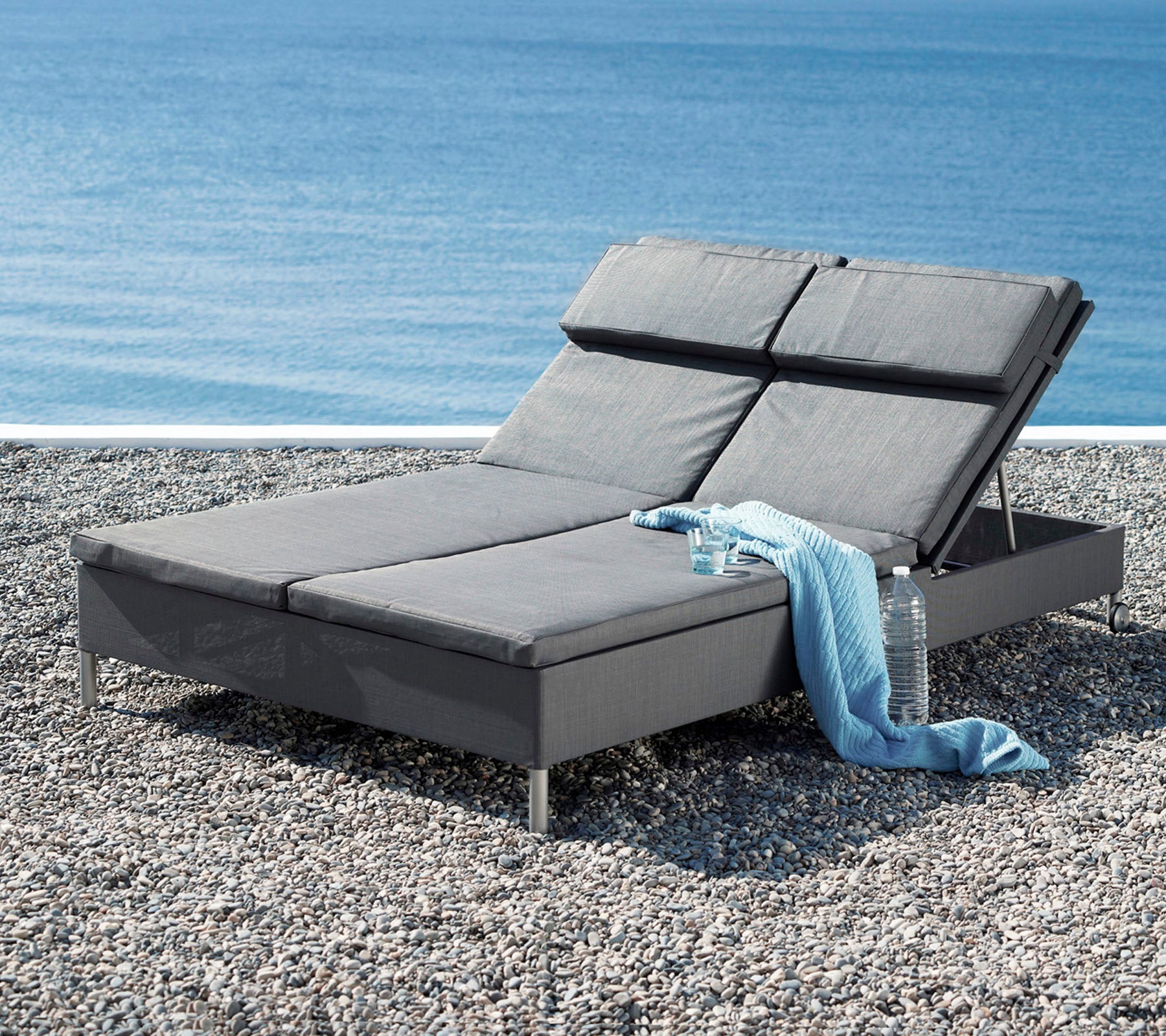 A stylish gray lounge chair with an adjustable backrest, accompanied by a soft throw, alongside a tranquil water view.