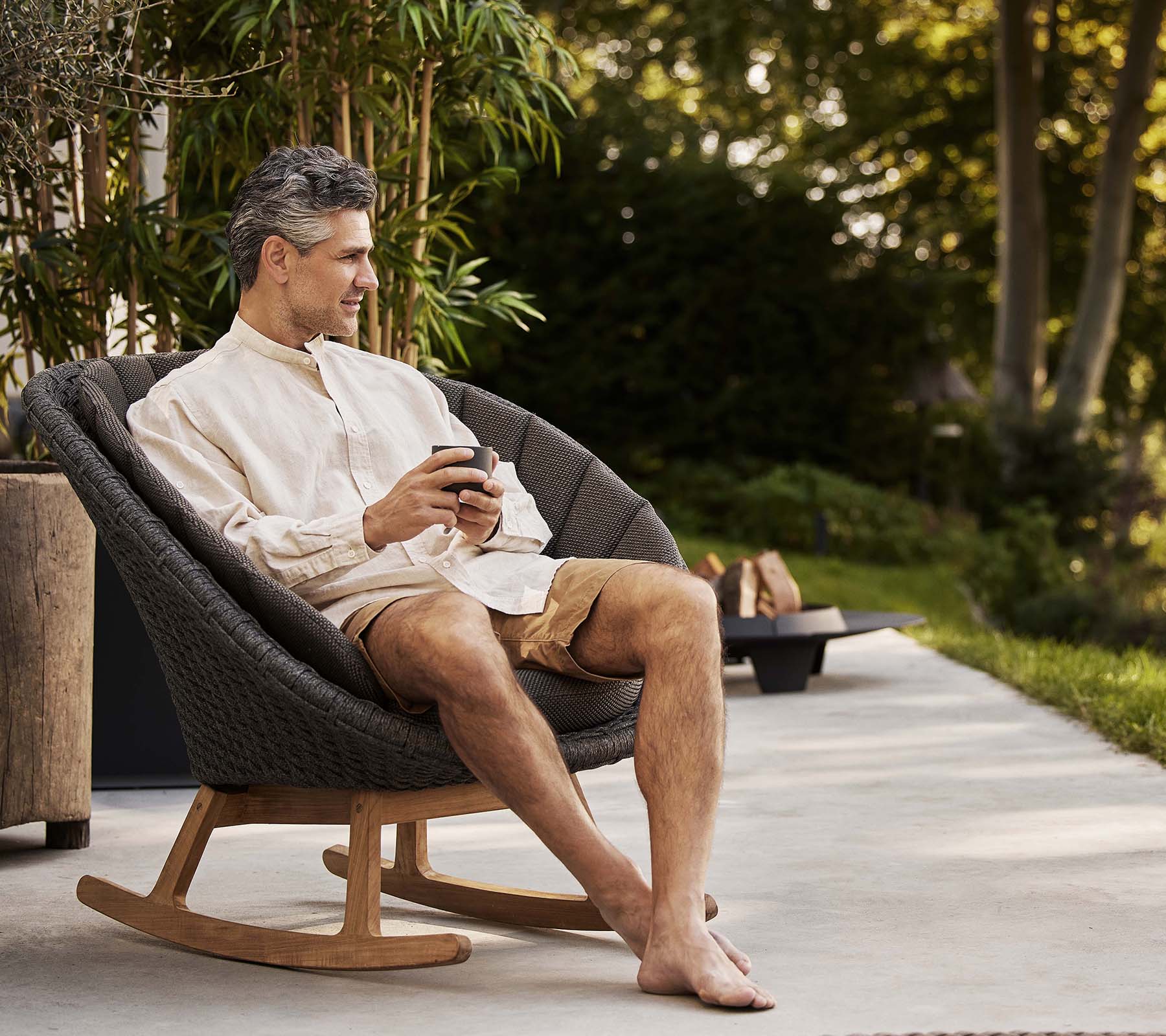 A man sits comfortably in a stylish peacock rocking chair, enjoying a moment of relaxation while holding a phone. Greenery is in the background.