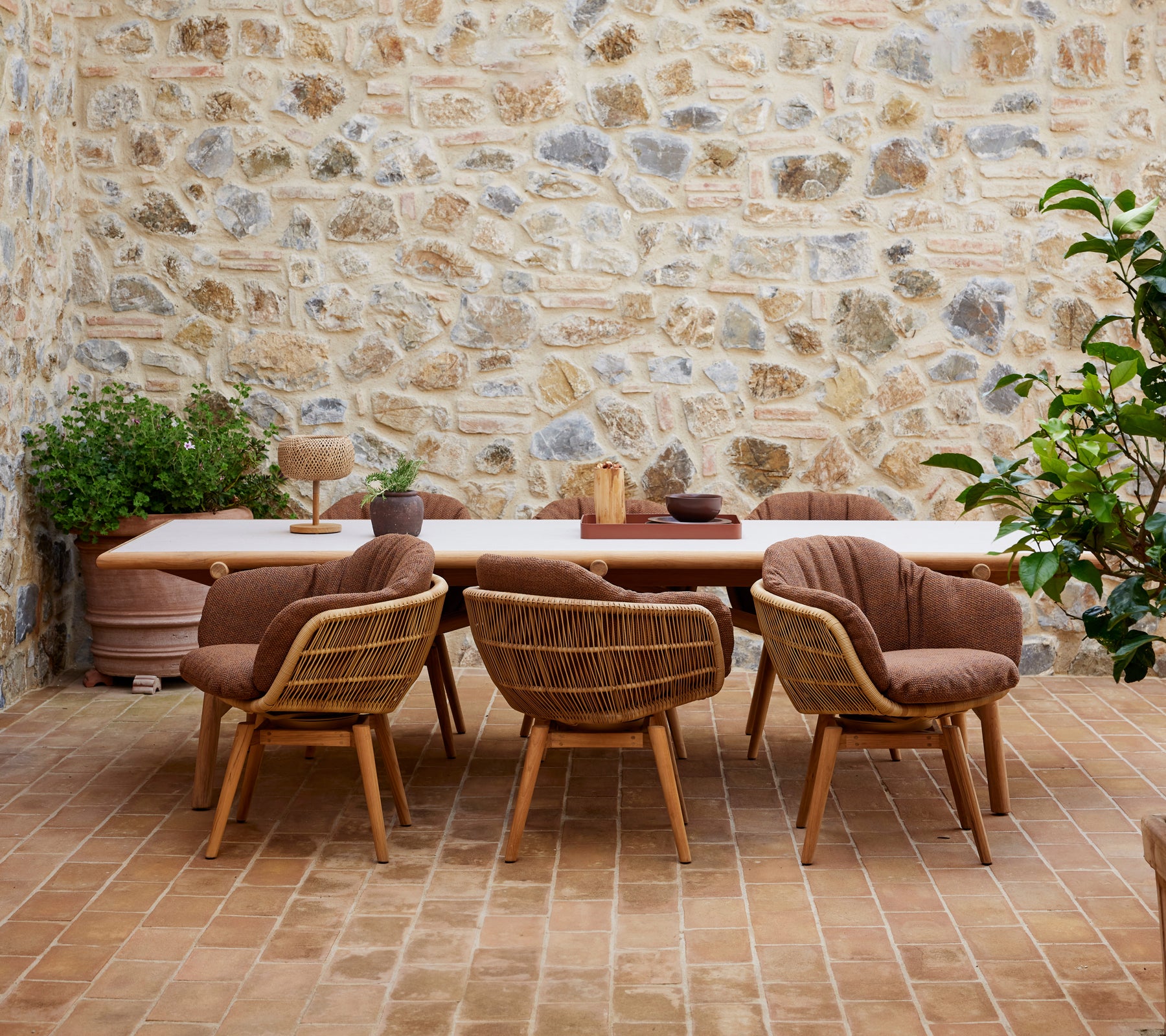 A wooden dining table surrounded by stylish chairs with teak legs, set against a textured stone wall and adorned with green plants.